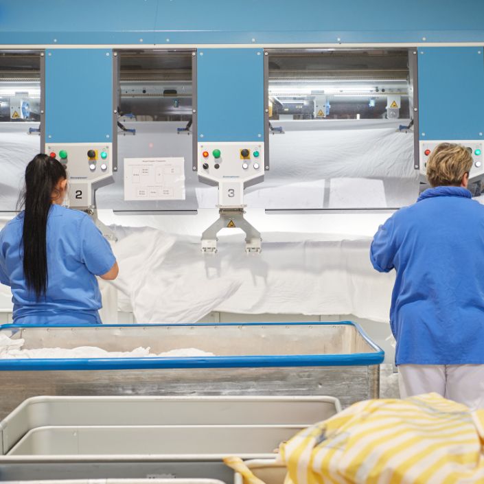 Zwei Wäscherei Mitarbeiterinnen bearbeiten weiße Mietwäsche an der Eingabemaschine einer Hightech Mangel. Zwischen ihnen ist ein Arbeitsplatz frei als Symbol für die freien Stellen in der Wäscherei.