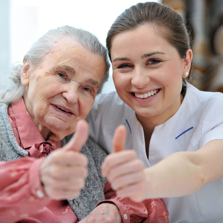 Eine Seniorin und eine weibliche Pflegekraft im Kassack strecken ihre Daumen nach oben