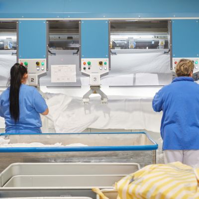 Zwei Wäscherei Mitarbeiterinnen bearbeiten weiße Mietwäsche an der Eingabemaschine einer Hightech Mangel. Zwischen ihnen ist ein Arbeitsplatz frei als Symbol für die freien Stellen in der Wäscherei.